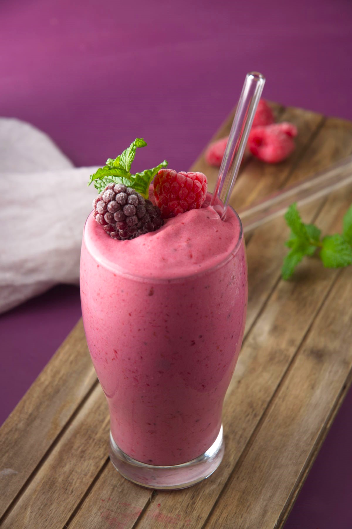 Triple berry smoothie in a glass with a clear straw on purple background.