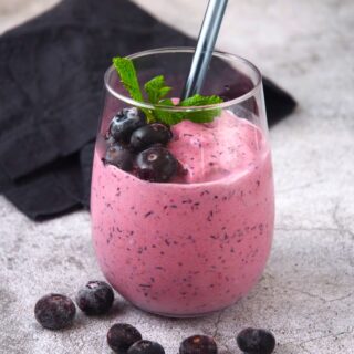 Keto blueberry smoothie in a stemless wine glass with straw, mint and fresh blueberries.