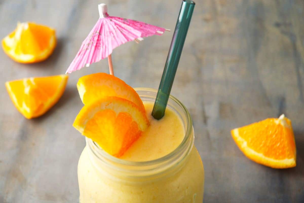 Mango pineapple smooth in glass jar with blue clear straw, orange slices and mini umbrella.