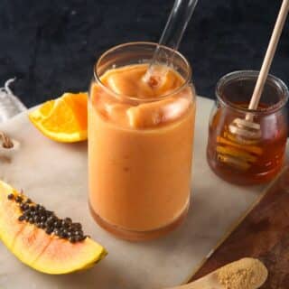 Papaya smoothie in a clear glass with clear straw and fresh papaya on the side.