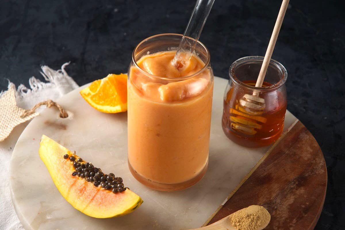 Papaya smoothie in a clear glass with clear straw and fresh papaya on the side.
