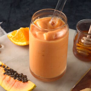 Papaya smoothie in clear glass with clear straw and fresh papaya on the side.