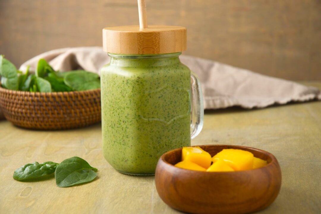 A green smoothie in a mason jar with a bamboo lid and straw sits on a wooden table. Next to it, there is a small wooden bowl containing mango chunks and a woven basket holding spinach leaves. A beige cloth is in the background.