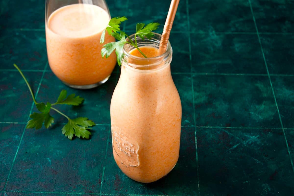 Carrot smoothie in glass with fresh parsley and straw on teal background.