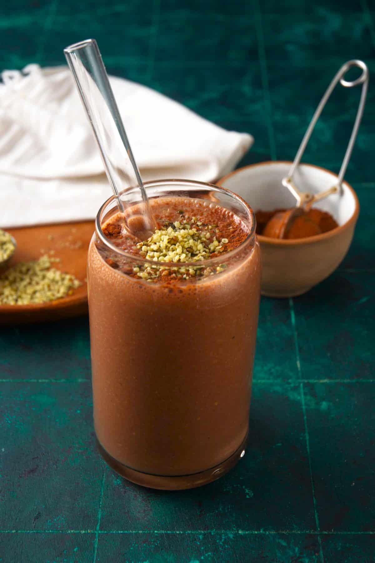 Chocolate peanut butter smoothie in glass with straw and topped with hemp seeds and cocoa powder on blue background.